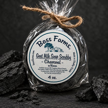Load image into Gallery viewer, A 4 oz round bar of Bass Farms Goat Milk Soap Scrubby in Charcoal, wrapped in clear cellophane and tied with twine, set on a dark textured surface with charcoal pieces.