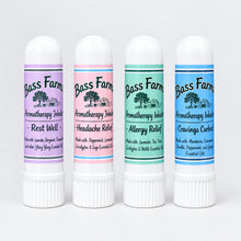 Load image into Gallery viewer, Four white lip balm tubes standing upright on a tan textured surface against a dark brown wooden background. Each tube has a different pastel label: lavender, pink, mint green, and blue, with the brand name 'Bass Farm' and product names 'Rest Well', 'Headache Relief', 'Allergy Relief', and 'Chronic Pain' printed on them.