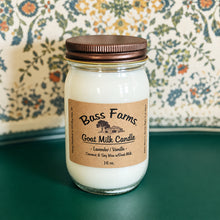 Load image into Gallery viewer, A white candle in a glass jar with a brown lid, labeled 'Bass Farms Goat Milk Candle, Lavender Vanilla' placed on a green surface with a patterned background.