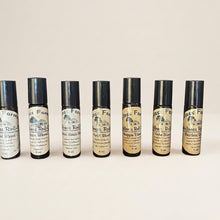 Load image into Gallery viewer, A row of roll-on amber glass bottles with black caps, displayed on a wooden shelf, containing various essential oil blends.